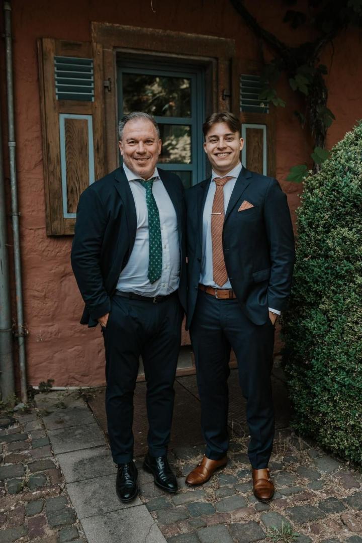 Zwei Männer in dunklen Anzügen stehen vor einem Gebäude mit Holztür.
